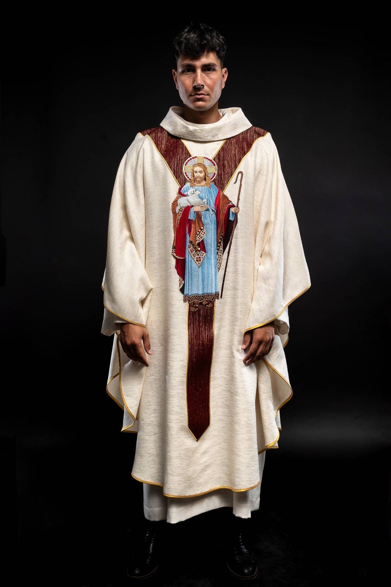 Casulla de color crudo con la imagen de Jesús Buen Pastor con una franja decorativa de color burdeos.